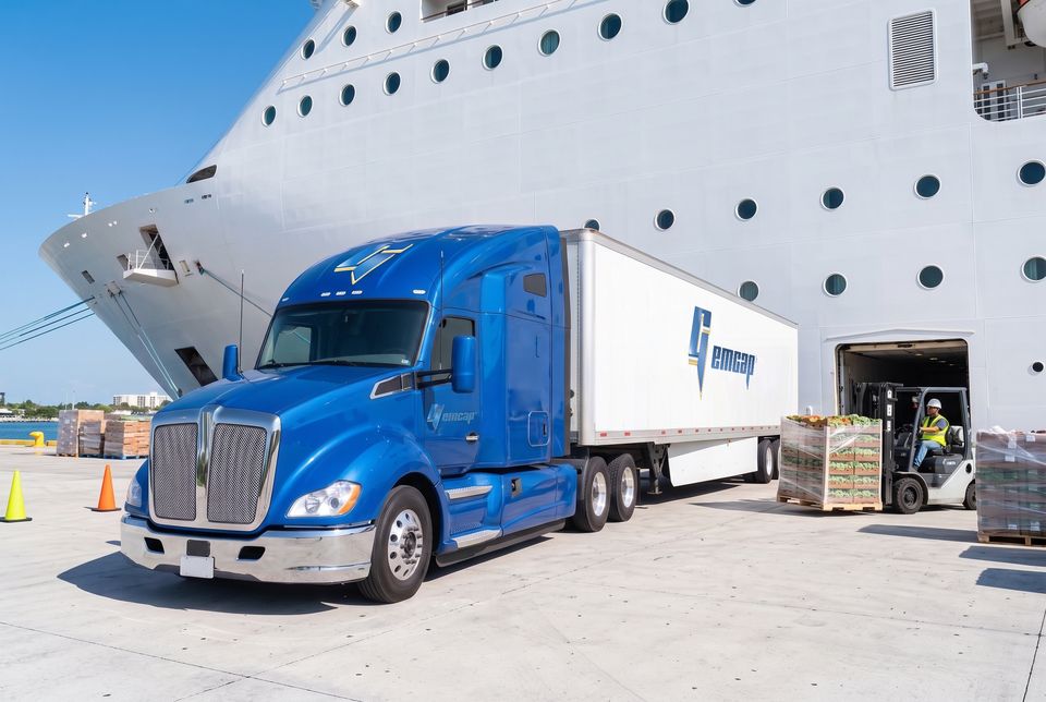 A blue GEMCAP tractor-trailer at the cruise pier while a forklift carries pallets of fresh produce into the ship