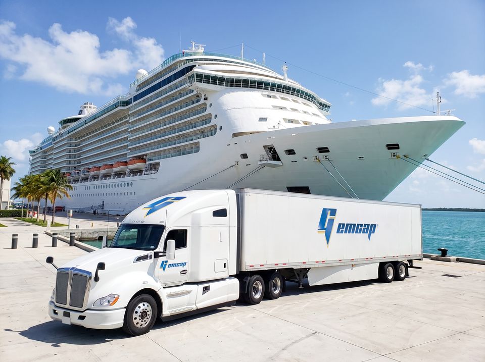 A white GEMCAP tractor-trailer parked on the pier at PortMiami beside a docked cruise ship