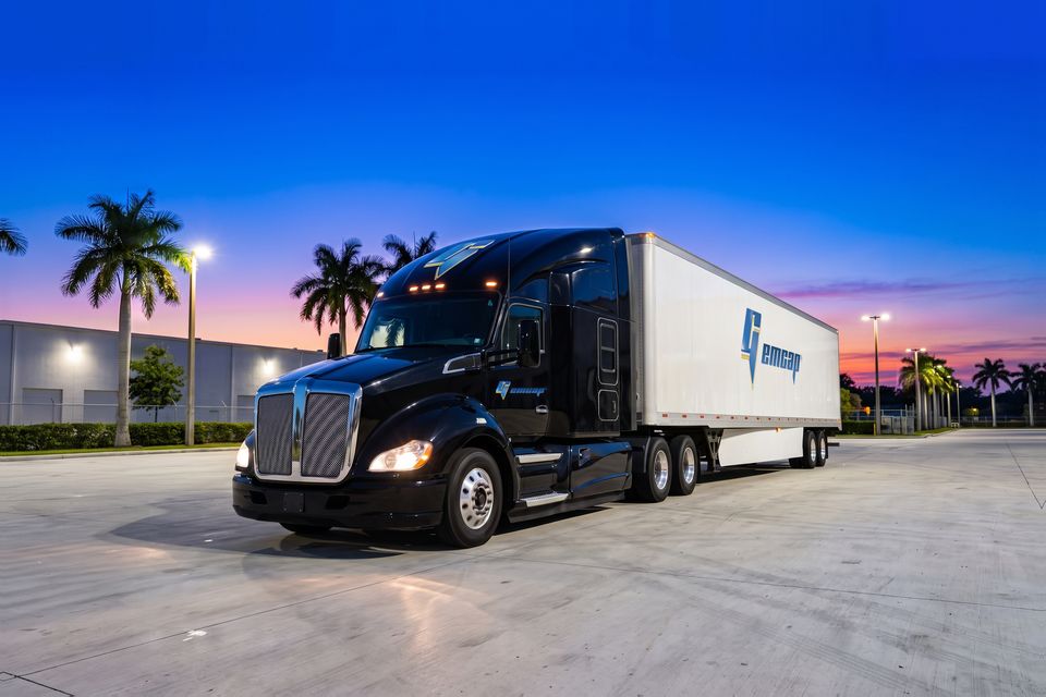 A black GEMCAP tractor-trailer with its headlights on in the Miami truck yard at blue hour